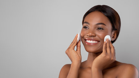 Woman Applying Skincare Product on Her Face and Smiling in a Bright Room With Neutral Backgroundの写真素材