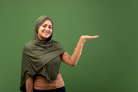 Cheerful Arab Lady in Hijab Pointing to the Side on Dark Green Backgroundの写真素材