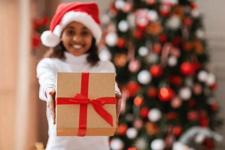 Happy Black Girl in Santa Hat Sharing Christmas Gift in Festive Living Roomの写真素材
