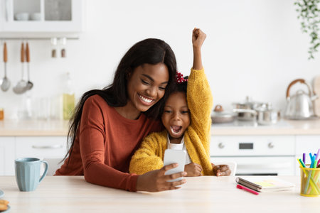 Excitement and Joy as Mother and Daughter Share a Special Moment Together While Using a Smartphone in the Kitchenの写真素材