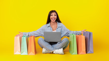 Joyful Shopper Sitting on the Floor With Shopping Bags and a Laptop in Front of a Bright Yellow Backgroundの写真素材