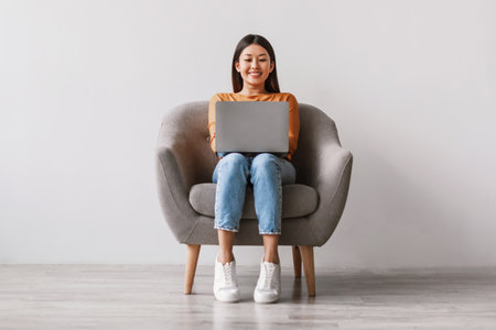 Young Asian Woman Working on Laptop in Cozy Armchair Against White Wallの写真素材
