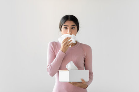 Young Woman Experiences Flu Symptoms While Holding Tissue Box Against Gray Wallの写真素材