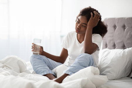 Young African American Lady Feeling Unwell in Bed While Holding Water Glass in Morning Lightの写真素材