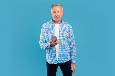 Mature Man Holds Smartphone While Standing in Blue Studio Backgroundの写真素材