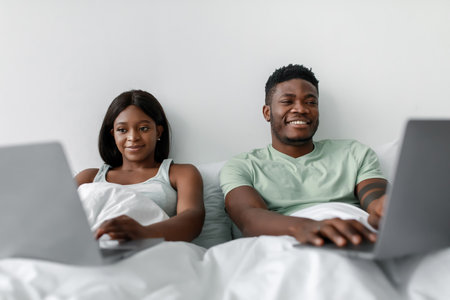 Couple Working on Laptops While Relaxing in Bed During a Cozy Indoor Eveningの写真素材