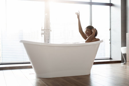 Positive Young Black Woman Enjoys Relaxing Bath While Listening to Music at Homeの写真素材