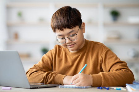 Plump Boy Studying at Home While Using Laptop and Taking Notes in Notebookの写真素材