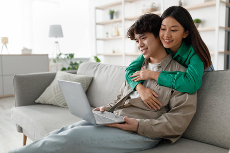 Young Asian Couple Enjoys Leisure Time at Home With Laptop and Affectionの写真素材