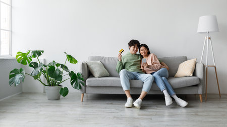 Young Asian Couple Enjoys Online Shopping Together on Mobile at Homeの写真素材