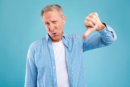 Senior Man Shows Thumbs Down Gesture, Expressing Disapproval in Blue Background Setting