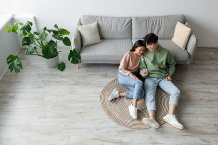 Cheery Asian Couple Relaxing at Home, Browsing Smartphone on Weekendの写真素材