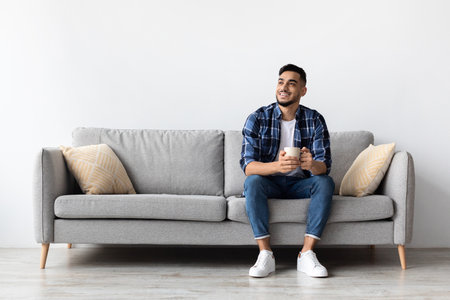 Happy Arab Man Relaxing at Home on Couch While Enjoying Coffee During Weekend Free Timeの写真素材