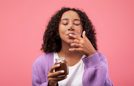Black Woman Enjoys Chocolate Paste From Jar, Licking Fingers on Pink Backgroundの写真素材