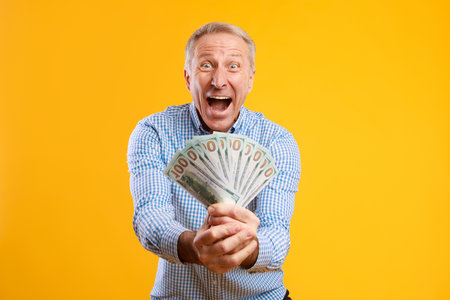 Excited Mature Man Shows off Cash in Hands Against Bright Yellow Backgroundの写真素材