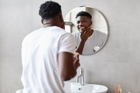 Happy Black Man Applying Moisturizer in Modern Bathroom for Facial Care Routineの写真素材