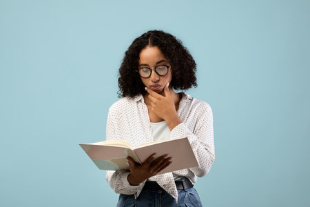 Pensive Black Female Student Reading Textbook While Studying for Exam Against Blue Backgroundの写真素材