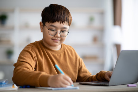 Intelligent Korean Kid Focuses on Homework at Home Using Laptop and Notebookの写真素材