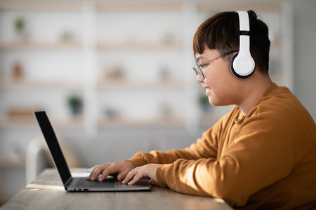Chubby Asian Teenager Using Laptop at Home While Smiling and Wearing a Wireless Headsetの写真素材