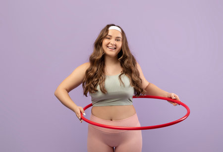 Smiling Young Plus Size Female in Uniform Exercises With Hula Hoop Against Purple Backgroundの写真素材
