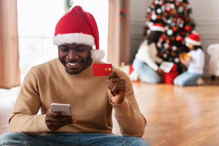 Happy Black Man Shopping Online for Christmas Gifts With Credit Card and Cellphone at Homeの写真素材