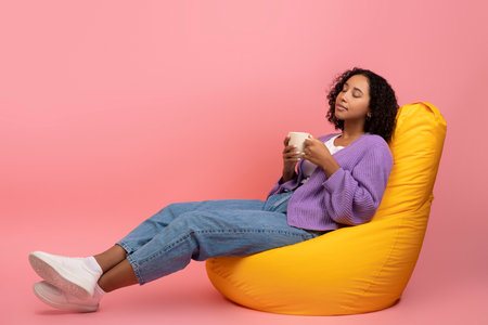 Millennial Black Female Relaxing in Bean Bag Chair With Coffee Against Pink Backgroundの写真素材