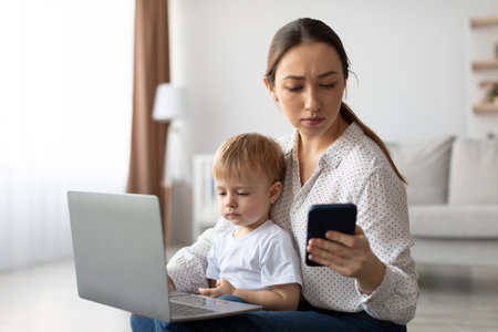 Mother Works on Laptop While Watching Toddler and Checking Phone in Cozy Living Roomの写真素材