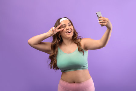 Cheerful Millennial Woman in Sportswear Taking Selfie and Livestreaming on Purple Backgroundの写真素材
