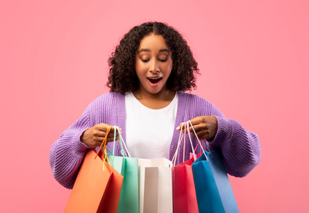 Young Black Woman Excitedly Explores Shopping Bags Against Pink Backgroundの写真素材