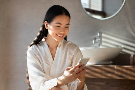 Cheerful Young Lady Enjoys Beauty Blog on Smartphone While Relaxing in Bathroomの写真素材