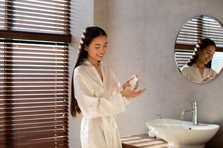 Long-haired Asian Woman Applies Beauty Product in Elegant Bathroom With Natural Lightの写真素材