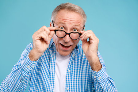 Surprised Mature Man in Glasses Reacts to Great Offer While Looking at Camera on Blue Backgroundの写真素材