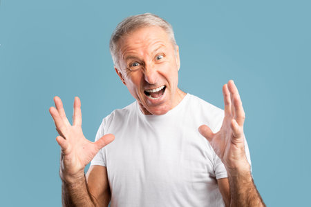 Mature Man Expressing Anger and Frustration While Standing Against a Blue Backgroundの写真素材