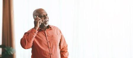 Elderly Man Smiling While Talking on the Phone by a Large Window in a Bright Roomの写真素材