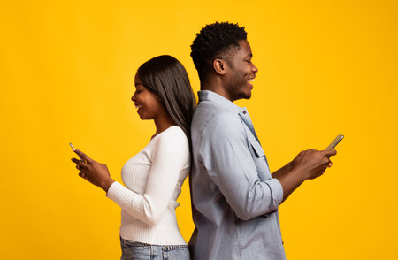 Friends Enjoying Their Smartphones While Standing Back to Back in a Bright Settingの写真素材
