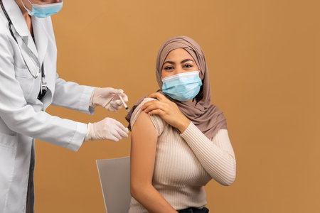 Doctor making injection of anti-covid vaccine to muslim woman in face mask and hijab, sitting over brown backgroundの写真素材