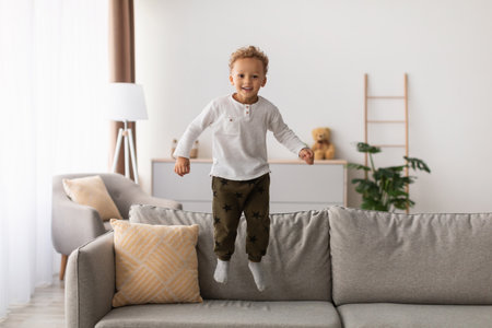 Cheerful Little Boy Jumping on the Couch at Home While Having Fun Indoorsの写真素材