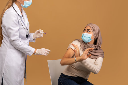 Young arab lady in face mask scared to make coronavirus vaccine injection, making STOP gesture and looking at doctorの写真素材
