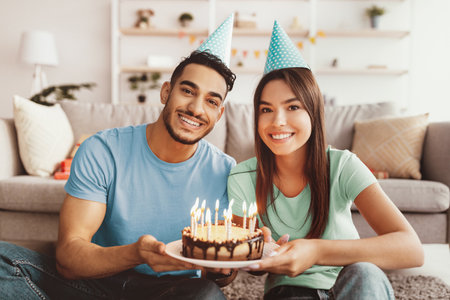 Happy Couple Celebrating Birthday With Cake and Party Hats in Cozy Living Roomの写真素材