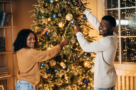 Young Black Couple Joyfully Decorating Christmas Tree at Home for Holiday Celebrationの写真素材