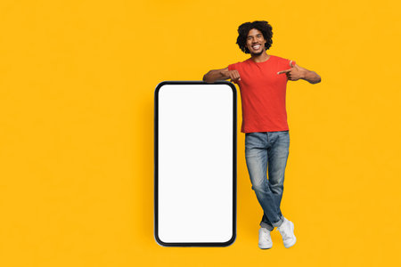 Smiling Man in Casual Outfit Poses Next to a Blank Smartphone on a Bright Yellow Backgroundの写真素材