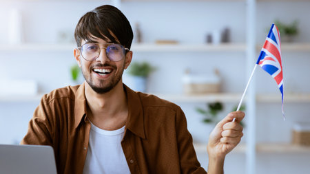 Young Man Celebrates With a Small British Flag While Sitting at His Desk With a Laptopの写真素材