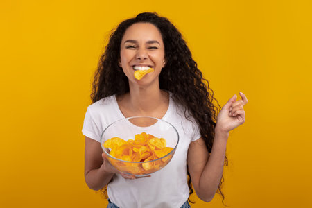 Excited Latin Woman Enjoying Tasty Potato Crisps in Cheerful Yellow Backgroundの写真素材
