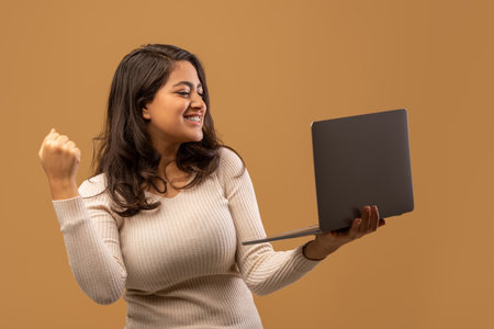 Overjoyed lady celebrating success with laptop, raising clenched fist, standing over beige backgroundの写真素材