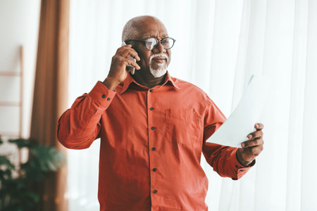 Elderly Man Talking on the Phone While Holding a Document in a Bright Indoor Settingの写真素材