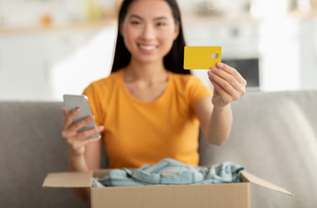 Korean Woman Enjoys Online Shopping Unpacking Clothes With Credit Card and Phone in Her Living Roomの写真素材