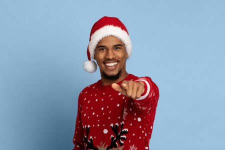 Man in Christmas Sweater and Hat Smiles While Pointing at the Camera Against a Blue Backgroundの写真素材