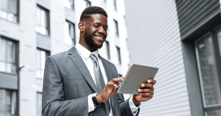 Professional Man Smiles While Using a Tablet Outside a Modern Office Buildingの写真素材