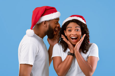 Man whispering christmas secret to surprised woman, spelling to her ear, wearing santa hats over yellow backgroundの写真素材