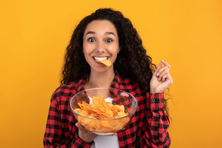 Latin Lady Joyfully Indulging in Crispy Potato Chips Against a Vibrant Yellow Backdrop